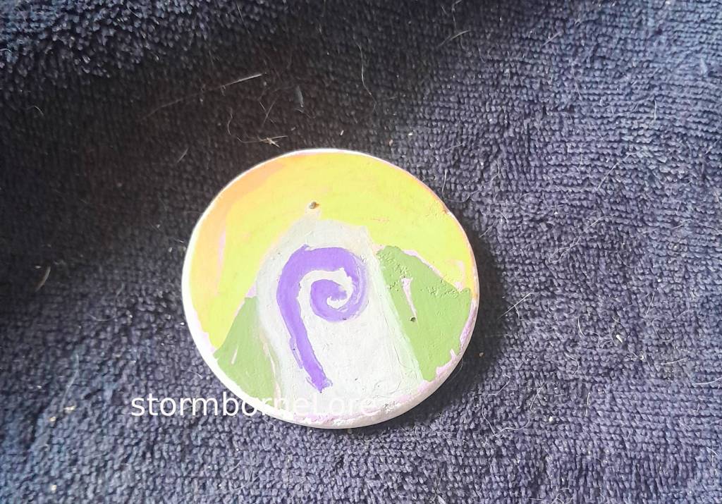 A round painted stone featuring an abstract mountain design with a spiral shape, placed on a textured dark fabric.