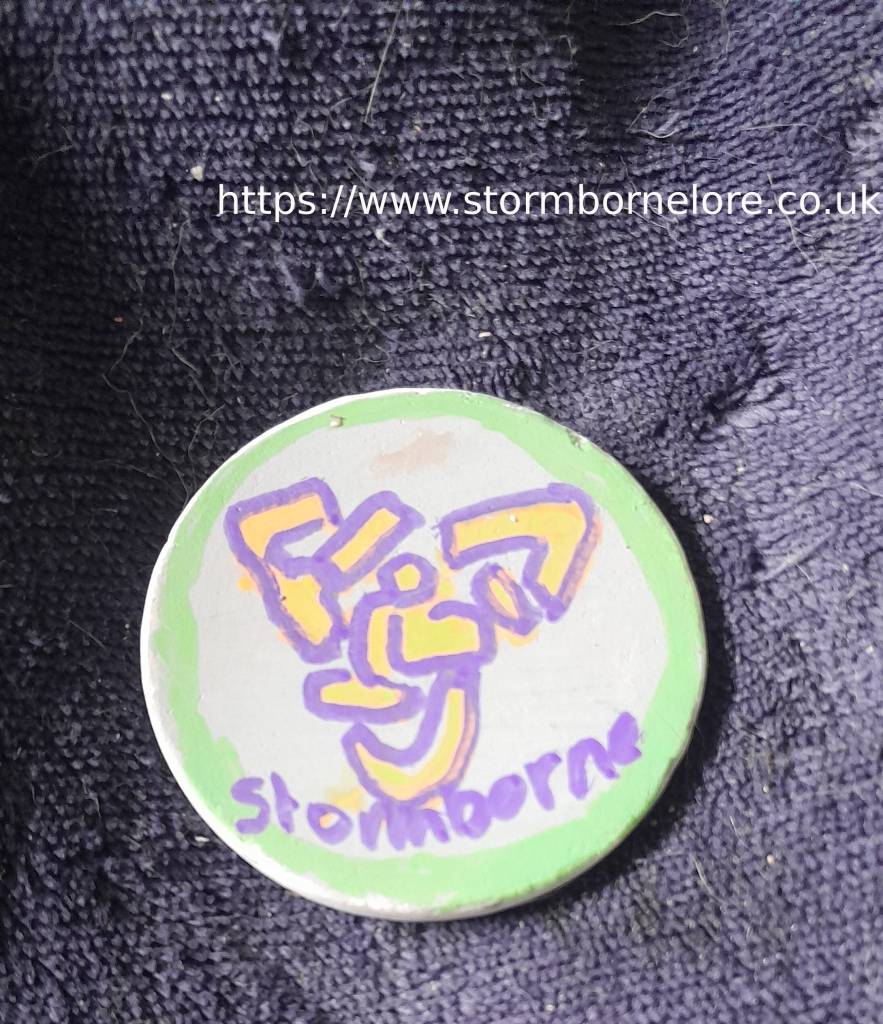 A round painted stone with abstract designs in purple and yellow on a gray background, encircled by a green rim, resting on a dark fabric surface.