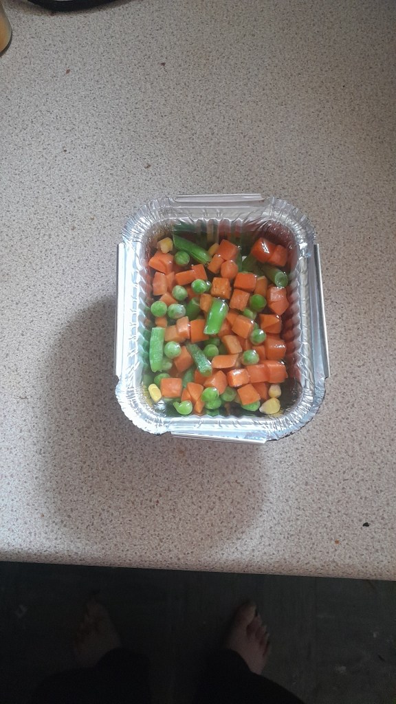 A foil tray filled with frozen mixed vegetables, including peas, carrots, green beans, and corn, placed on a kitchen countertop.