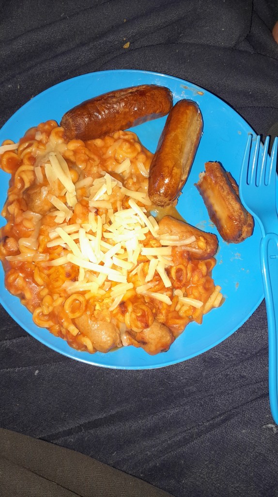 A blue plate with gluten-free pasta, baked beans, sausage slices, and a sprinkle of cheese, accompanied by blue cutlery.