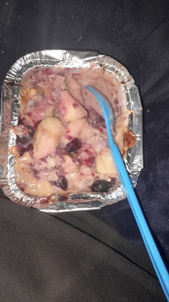 A small foil dish containing a creamy mixture of baked apples, blueberries, and raspberries, topped with custard and oats, with a blue spoon resting in it.