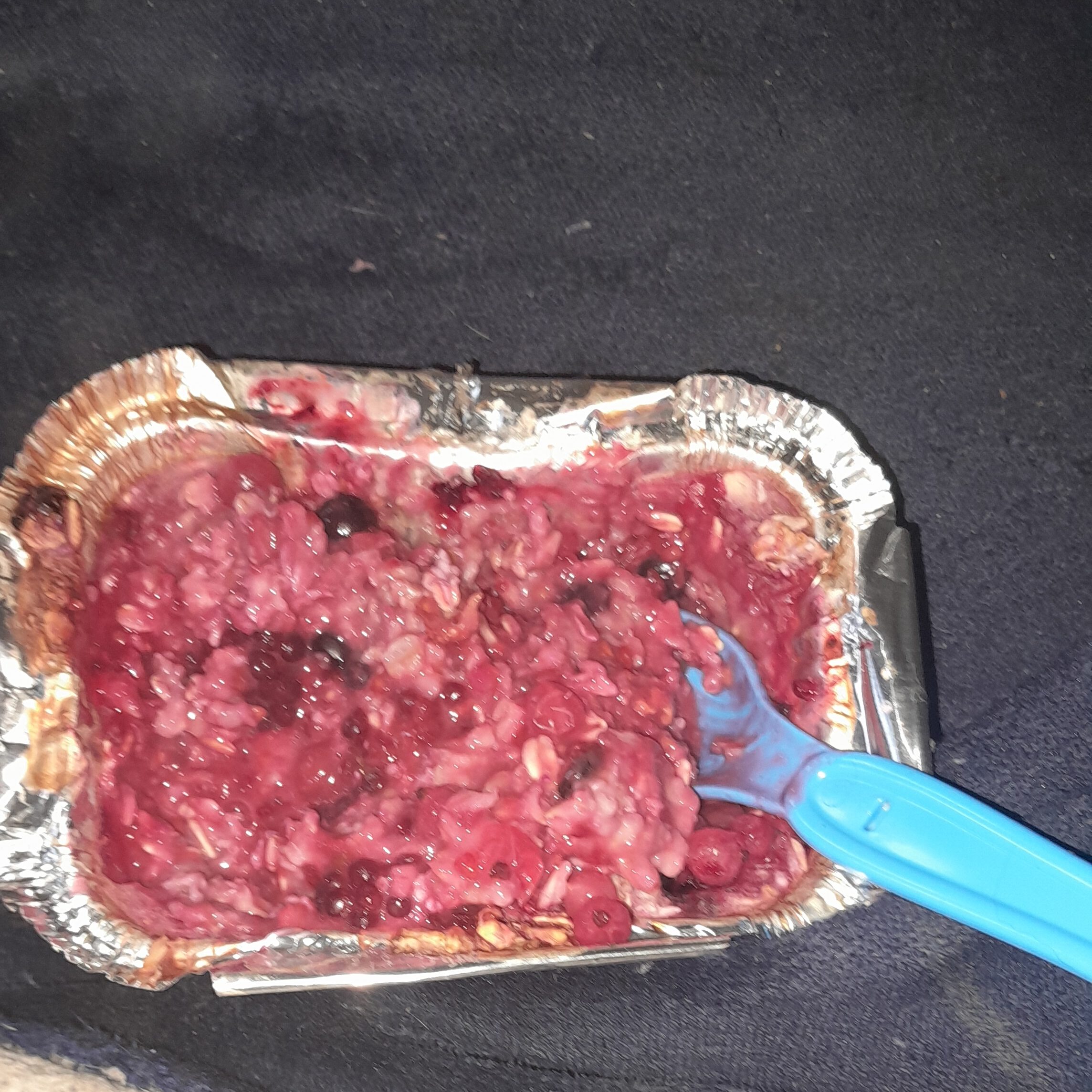 A small foil tray containing warm budget breakfast, fruity porridge made from oats and summer berries, with a blue spoon resting inside.