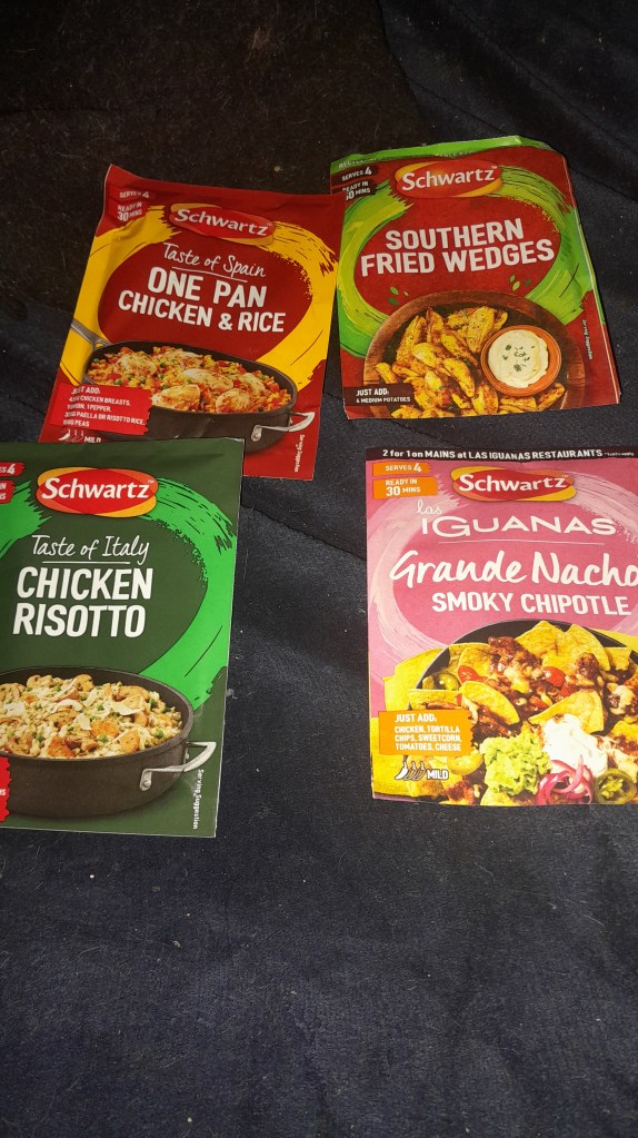 Four meal seasoning packs displayed on a black surface, including 'Taste of Spain One Pan Chicken & Rice,' 'Southern Fried Wedges,' 'Taste of Italy Chicken Risotto,' and 'Grande Nachos Smoky Chipotle' from Schwartz.