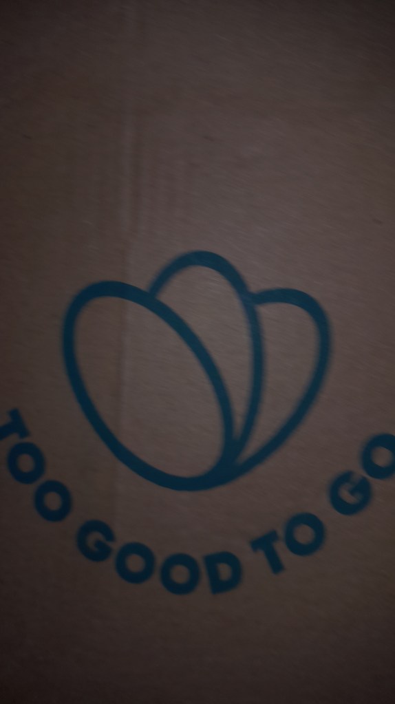 Close-up of the 'Too Good To Go' logo on a box made of brown cardboard, featuring two overlapping oval shapes and the text 'TOO GOOD TO GO' in a circular pattern.