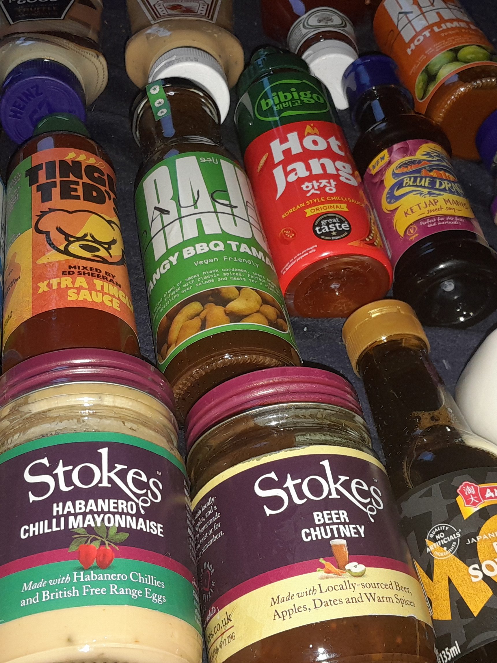 A variety of sauce bottles and jars, including Stokes Habanero Chilli Mayonnaise, Stokes Beer Chutney, Tingly Ted's Extra Tingly Sauce, Raju Tangy BBQ Tamarind, Bibigo Hot Jang Chilli Sauce, and Blue Dragon Ketjap Manis, arranged together on a surface.