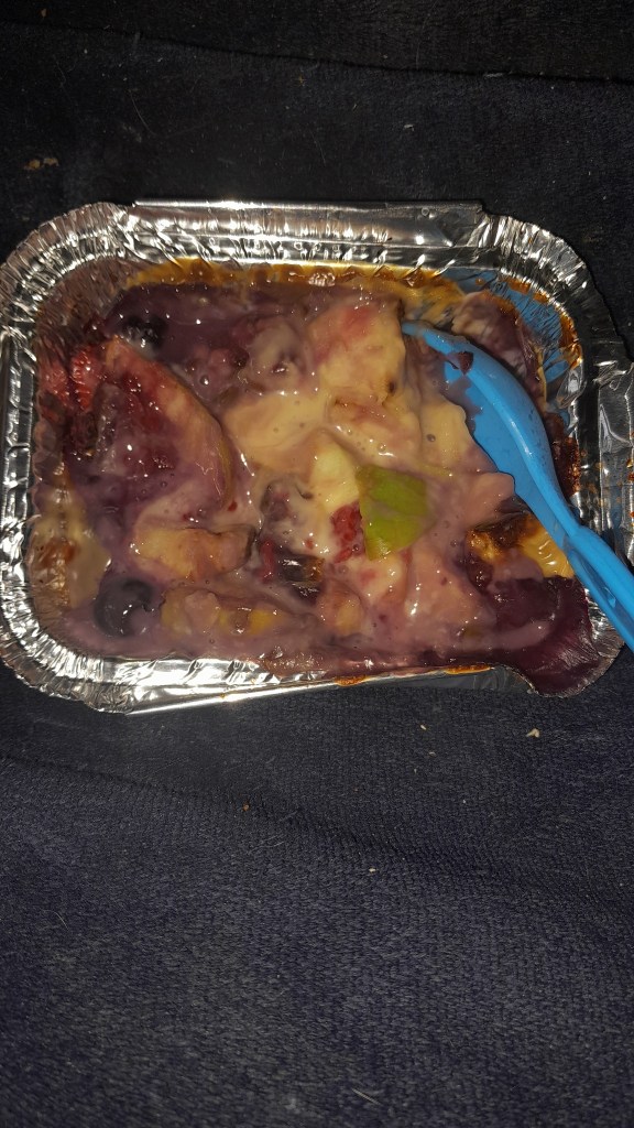 A foil container filled with a colorful dessert, featuring fruit mixed with a creamy purple sauce and topped with a light blue spoon.
